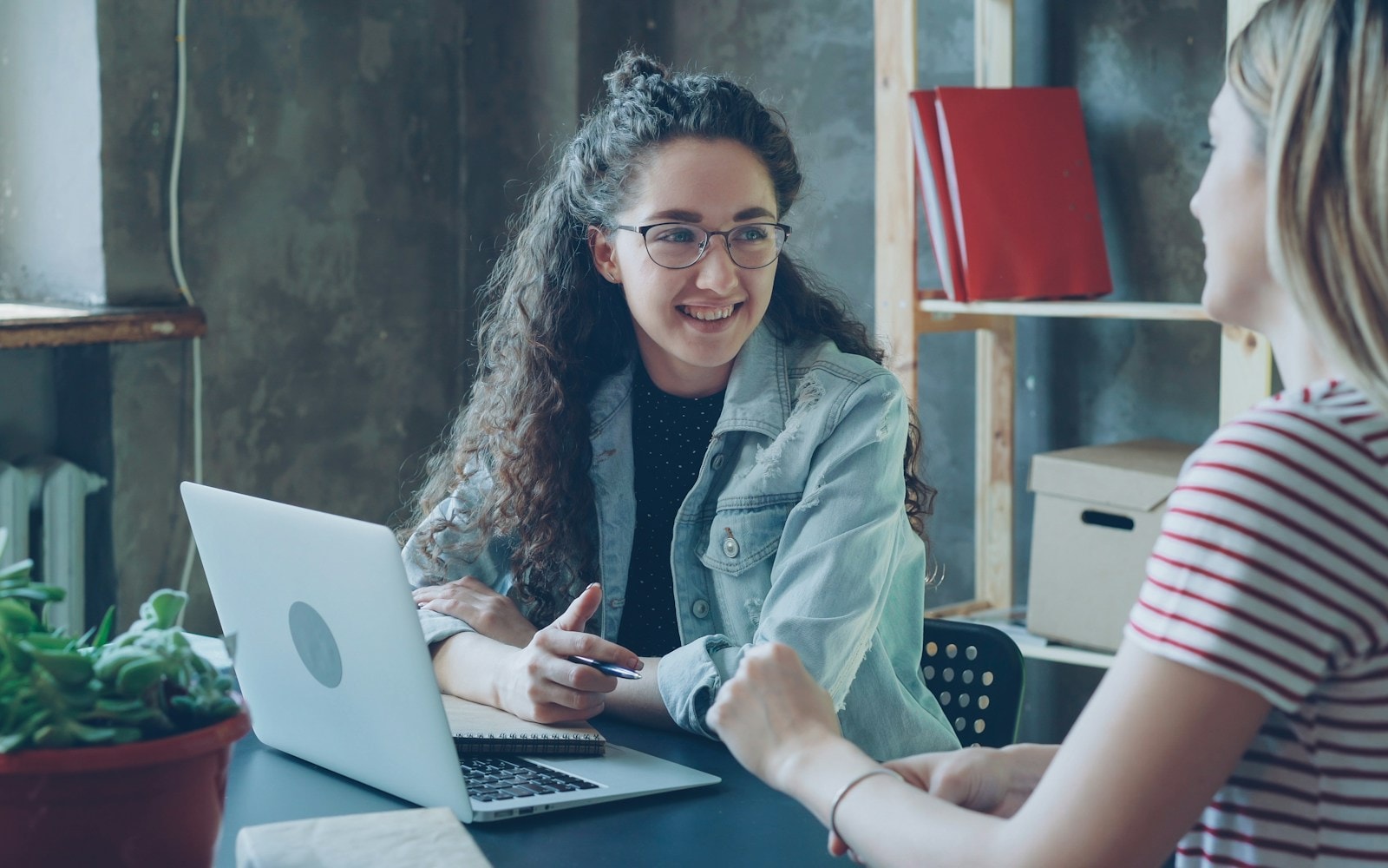 Dos personas conversando en una mesa de trabajo, con un computador portátil abierto.